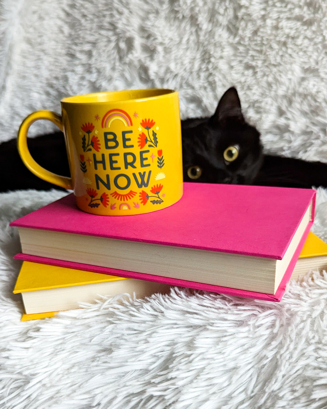 A yellow mug with florals and words "Be Here Now" sits on top of a pink hardcover book. A black cat lays behind and is peeking  with one eye.